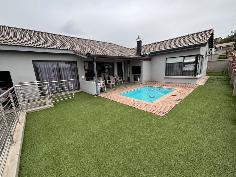 Rear exterior view featuring a small swimming pool, brick paved surround, artificial lawn, and a covered patio area with an external fireplace.