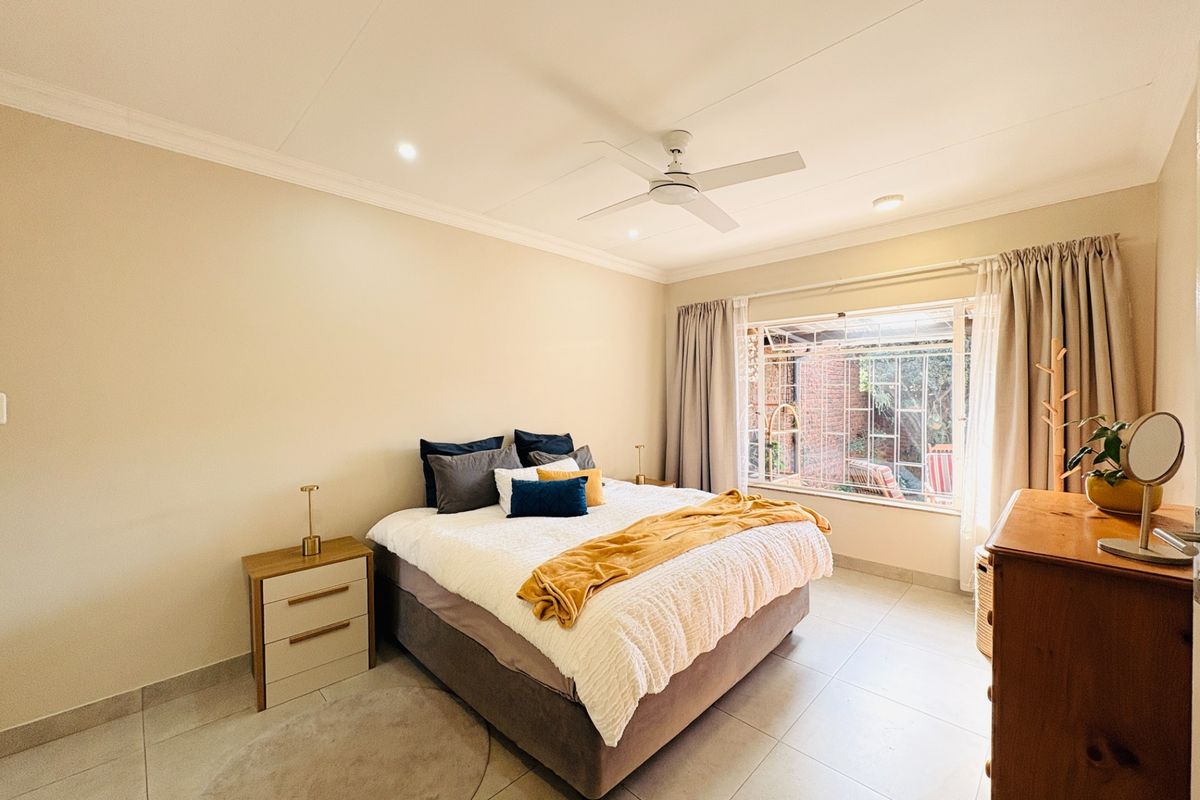 A well-lit bedroom featuring neutral tiled flooring, a ceiling fan, and a large window with security bars overlooking the rear exterior area.