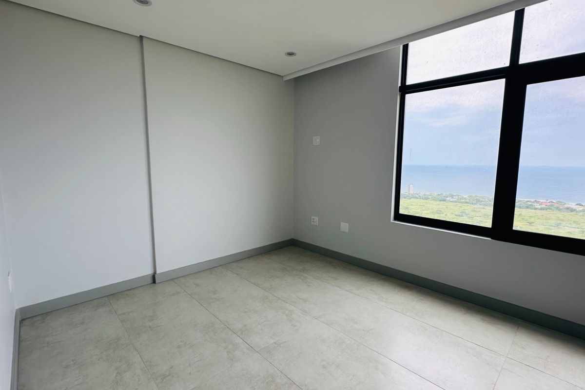A bright, empty bedroom featuring large windows with expansive coastal views, neutral tiled flooring, and modern recessed ceiling lighting.