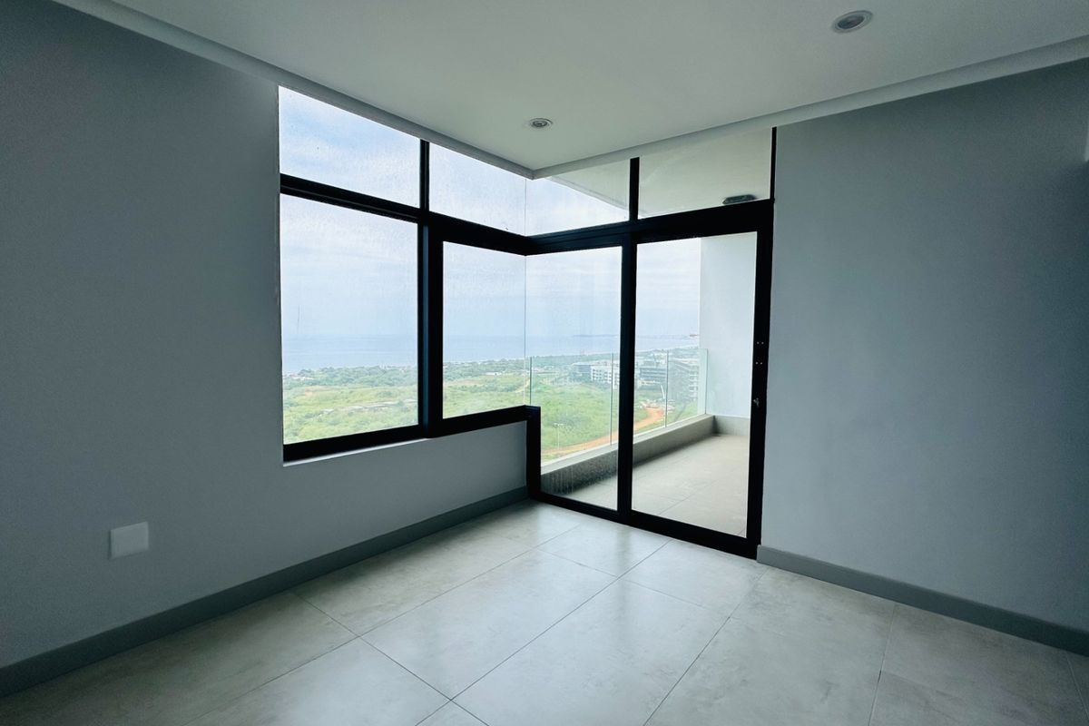 A bright, empty living area featuring large floor-to-ceiling windows and a glass door leading to a private balcony with scenic views.