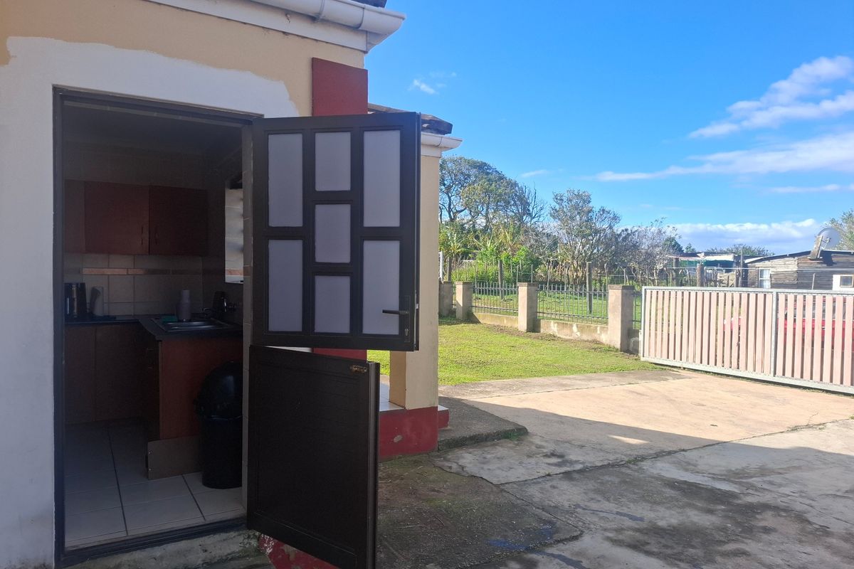 Rear view of the property showing a concrete driveway, metal gate, and access to a small kitchen area with cabinetry and tiled flooring.