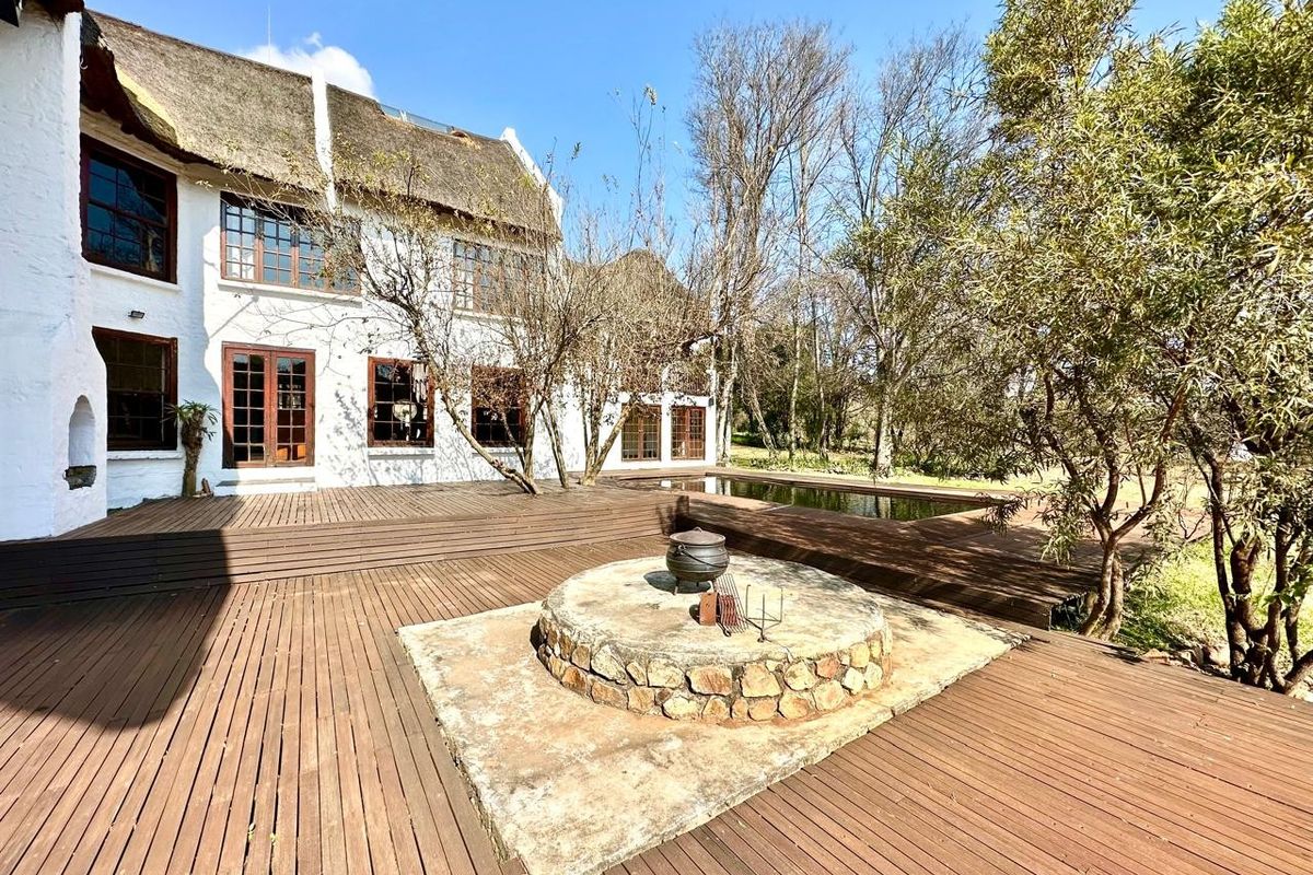 Rear exterior view featuring a large timber deck, stone fire pit, swimming pool, and a traditional thatched roof farmhouse facade.