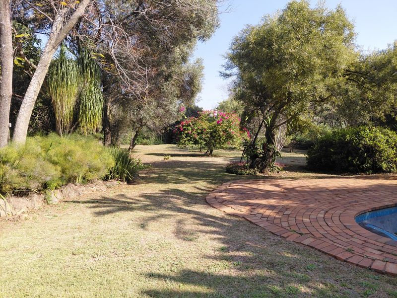 A spacious garden area featuring mature trees, established landscaping, and a brick-paved pool surround.