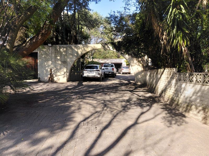 A gated entrance featuring a distinctive stone archway leading to a paved driveway and parking area surrounded by mature trees.