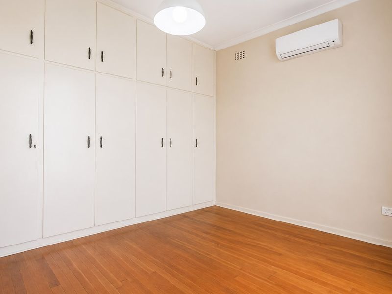 A clean, empty bedroom featuring polished timber flooring, extensive built-in cabinetry, and a wall-mounted split-system air conditioning unit.