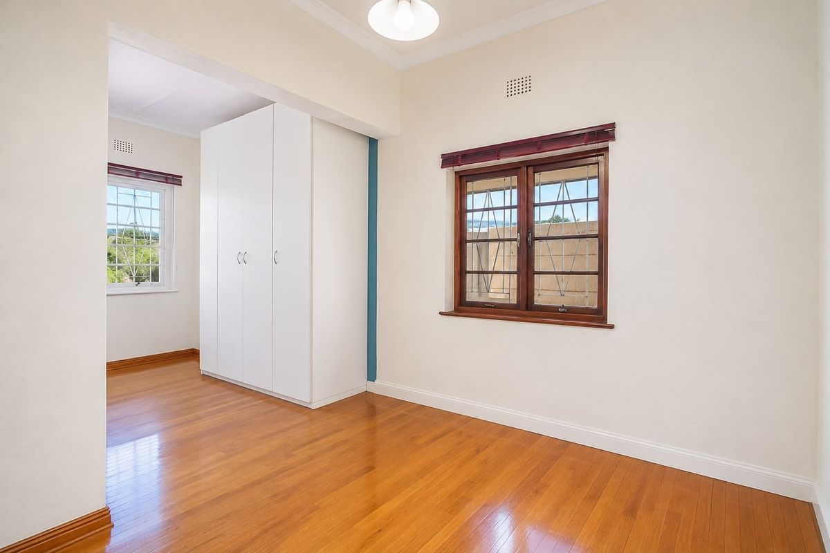 A bright, empty bedroom featuring polished timber flooring, built-in white cabinetry, and wooden-framed windows with security bars.