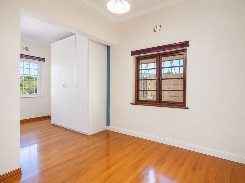 A bright, empty bedroom featuring polished timber flooring, built-in white cabinetry, and wooden-framed windows with security bars.
