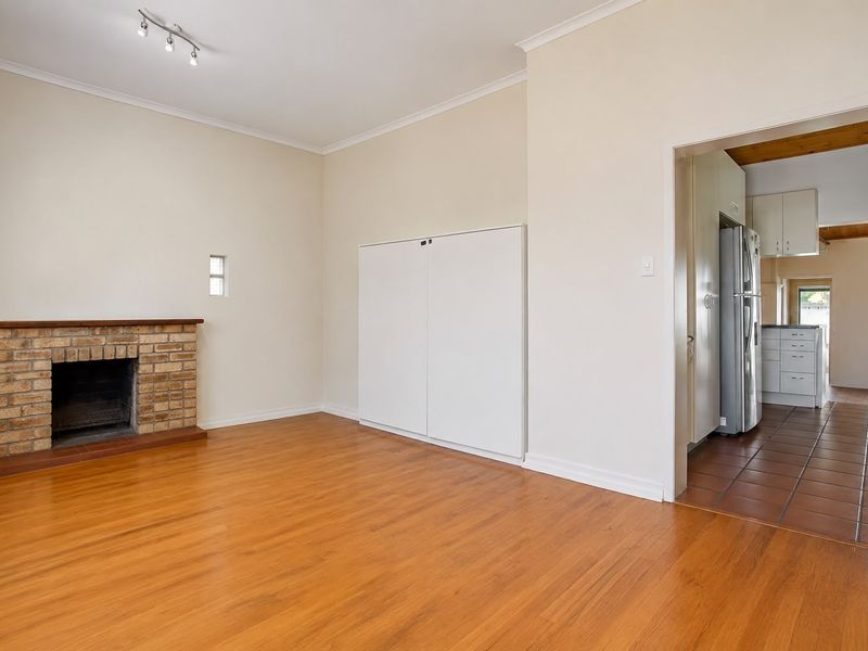 A bright living room featuring a brick fireplace and wood-look flooring, with an adjacent kitchen area visible through the doorway.