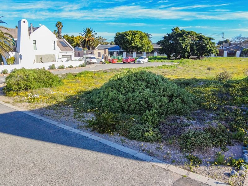 A vacant residential plot featuring natural vegetation, paved driveway access, and established kerbing in a suburban neighborhood setting.