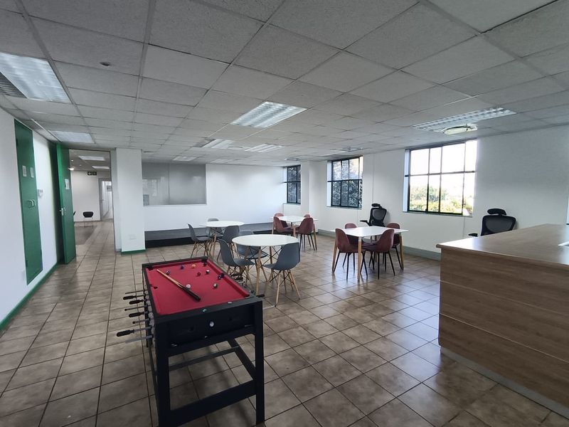 Open-plan office breakout area featuring tiled flooring, suspended ceiling grid, and large windows providing natural light.