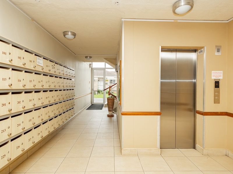 A clean, well-maintained retirement village hallway featuring a bank of built-in mailboxes, a stainless steel elevator, and tiled flooring leading to the main entrance.