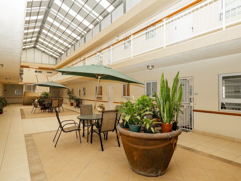 A bright, covered atrium area within the retirement village, featuring tiled flooring, natural light from the overhead roof, and access to residential units.