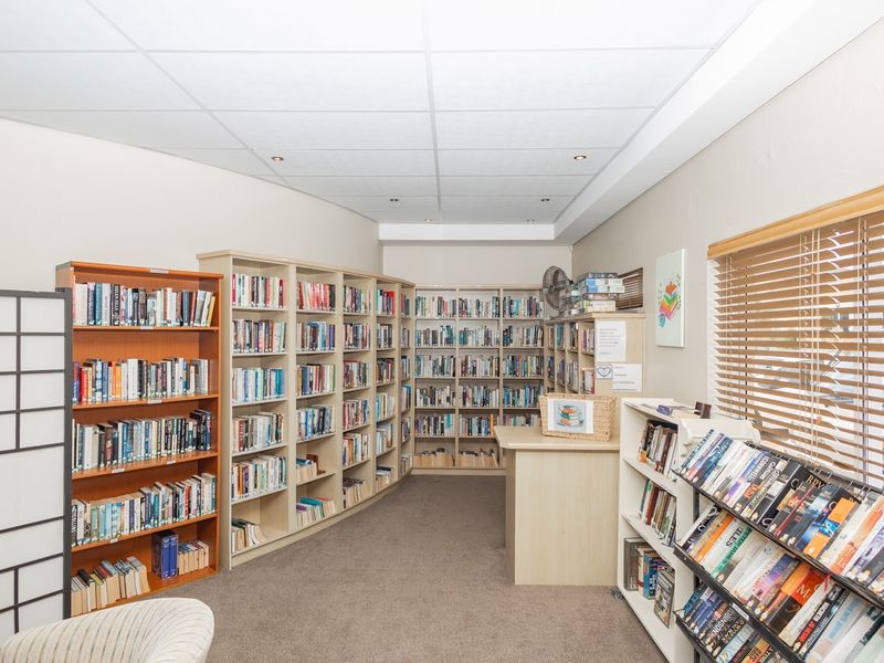 A communal library space featuring extensive built-in shelving units, recessed lighting, and large windows with venetian blinds.