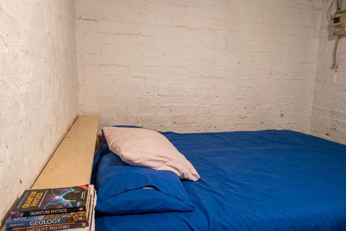 A compact bedroom featuring painted exposed brick walls and a simple built-in wooden headboard shelf.