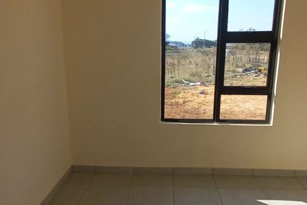 A bright, empty bedroom featuring neutral tiled flooring, cream-colored walls, and a dark-framed window overlooking the surrounding landscape.
