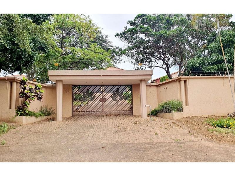 Front entrance view featuring a paved driveway, perimeter wall with electric fencing, and a secure metal gate under a covered structure.