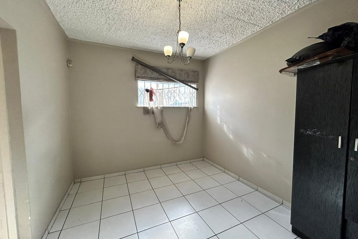A compact bedroom featuring white floor tiles, a textured ceiling, and a window fitted with security bars.