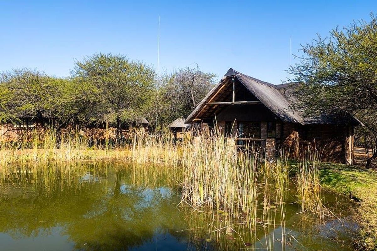 A scenic view of a rustic thatched-roof farm building situated beside a natural pond, surrounded by mature trees and native vegetation.