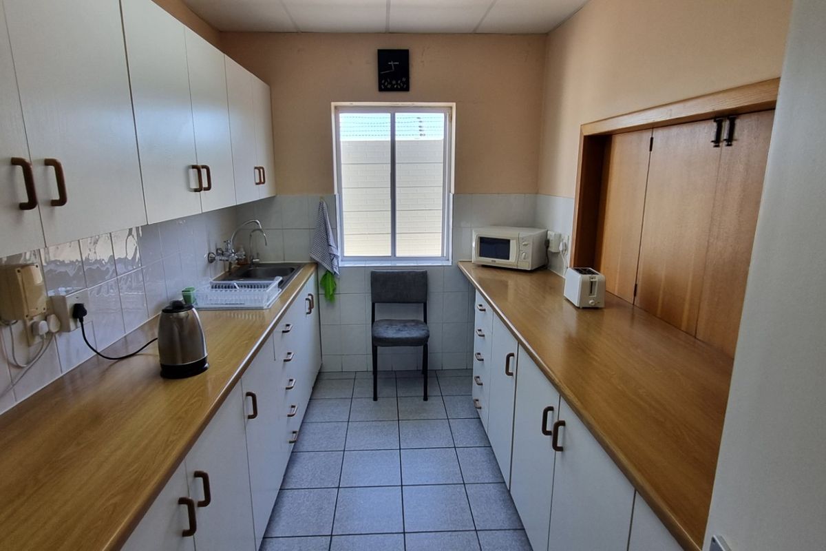 A functional galley-style office kitchenette featuring white cabinetry, wood-look laminate countertops, tiled flooring, and a stainless steel sink.