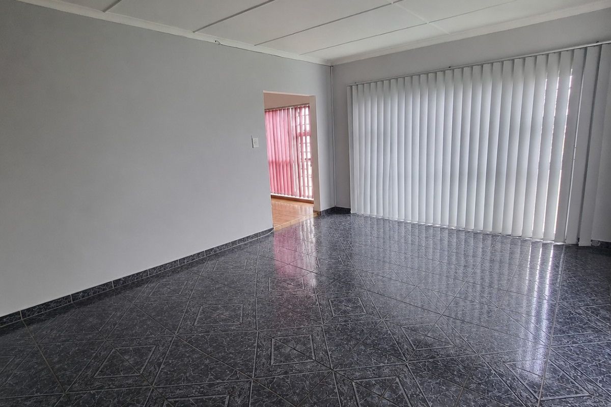 A spacious, empty living room featuring dark patterned floor tiles, neutral walls, and vertical blinds covering a large window.