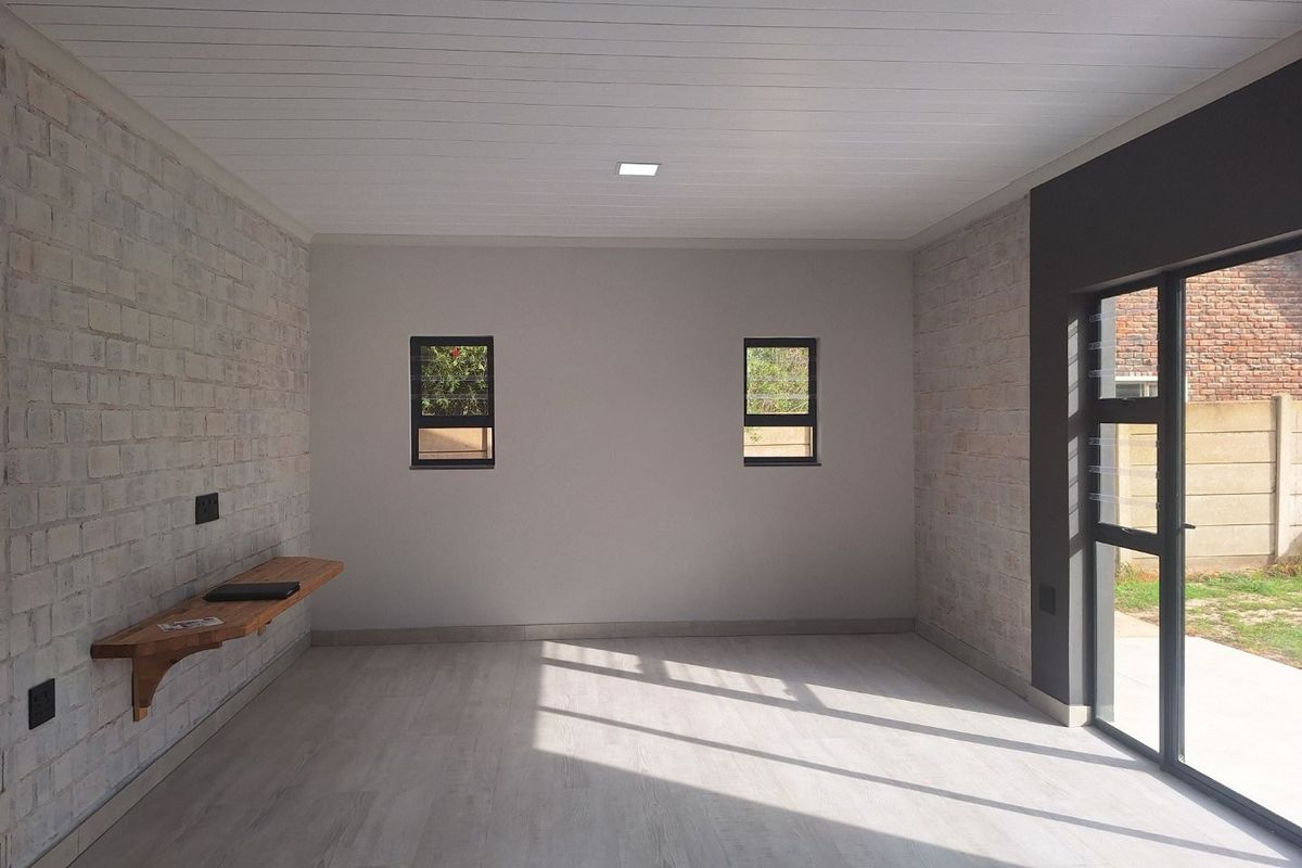 A bright, modern living area featuring exposed brick walls, light wood-look flooring, a timber wall-mounted shelf, and large glass sliding doors leading outdoors.