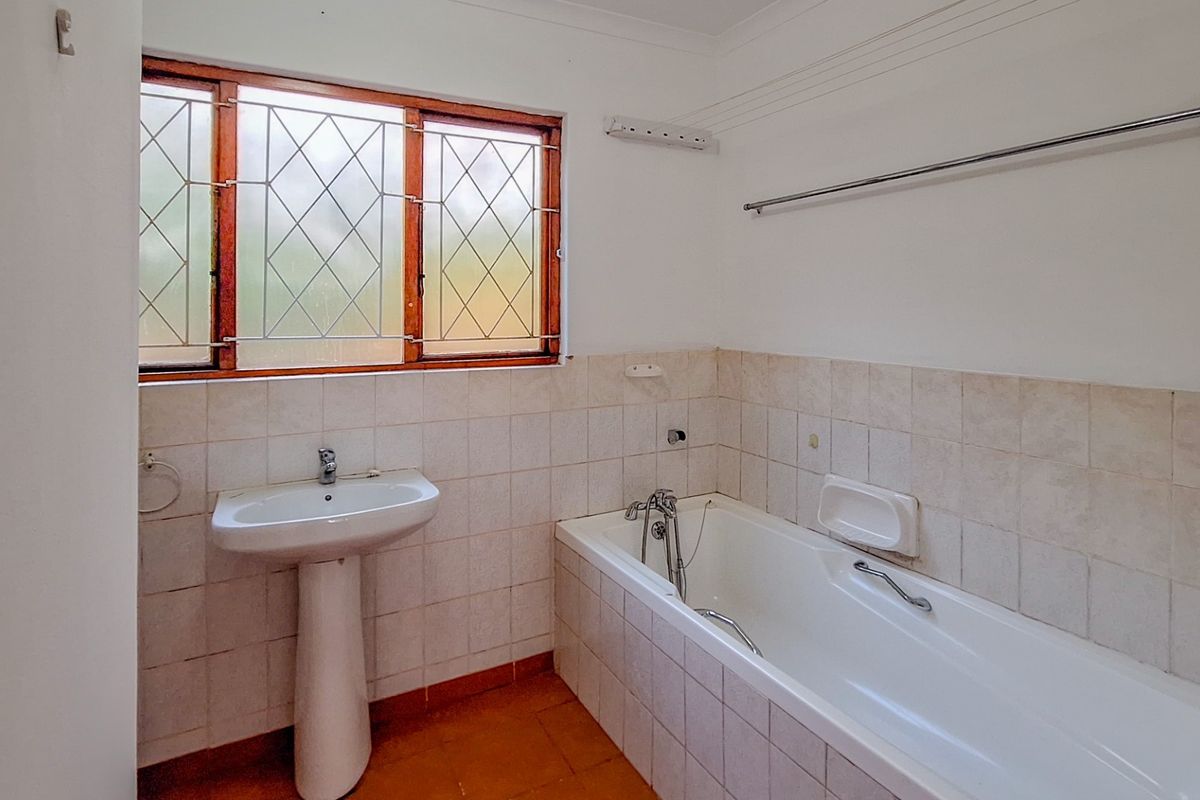 A functional bathroom featuring a pedestal basin, a built-in bathtub with tiled surround, and a window with security bars.