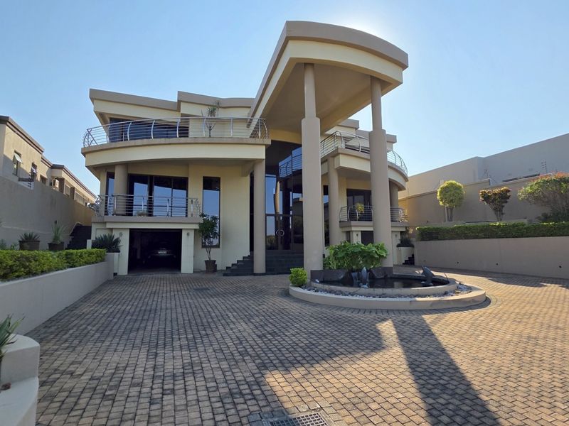 Modern multi-level architectural home featuring a paved driveway, integrated garage, circular water feature, and expansive balconies with stainless steel railings.