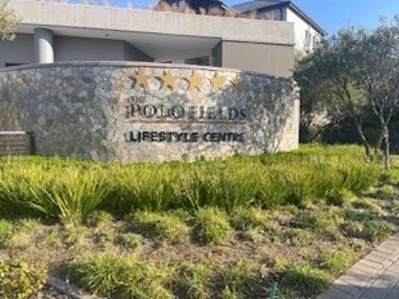 Entrance signage and landscaped garden area for The Polo Fields Lifestyle Centre, featuring stone cladding and paved pathways.