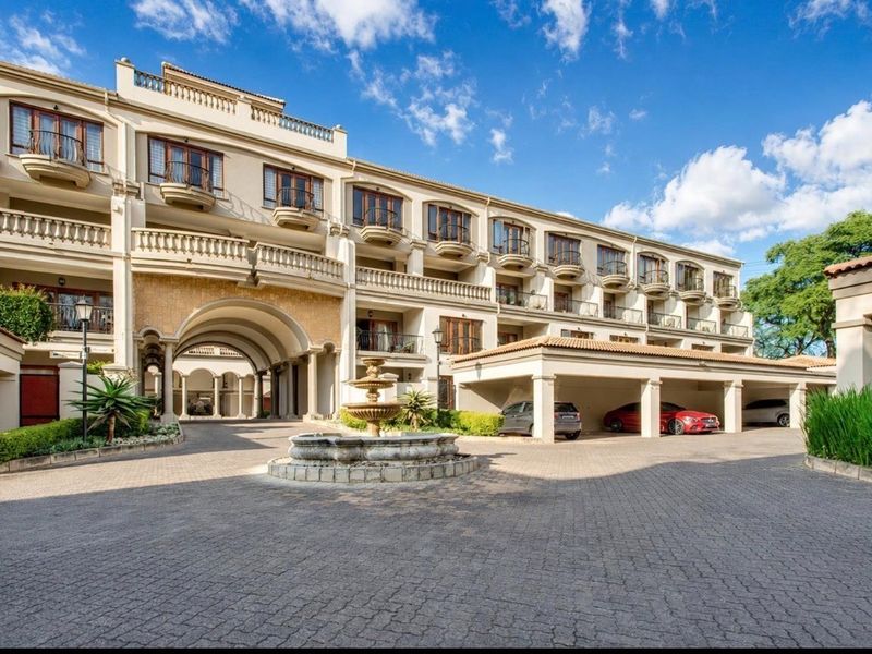 A view of the apartment complex exterior featuring a paved driveway, covered parking bays, and a central decorative fountain.