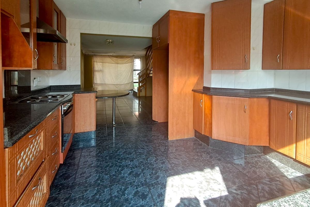 A functional kitchen featuring wood-toned cabinetry, dark granite countertops, and tiled flooring, leading into an open-plan living area with a staircase.