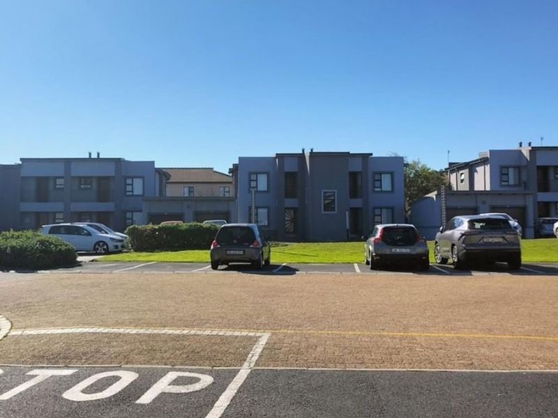 A view of the apartment complex exterior featuring a paved parking area, landscaped lawn, and secure perimeter fencing.
