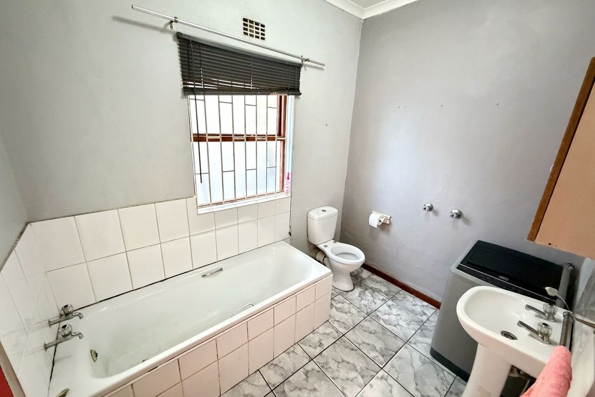 A functional bathroom featuring a tiled bathtub, pedestal basin, and toilet, with marble-effect floor tiles and security bars on the window.