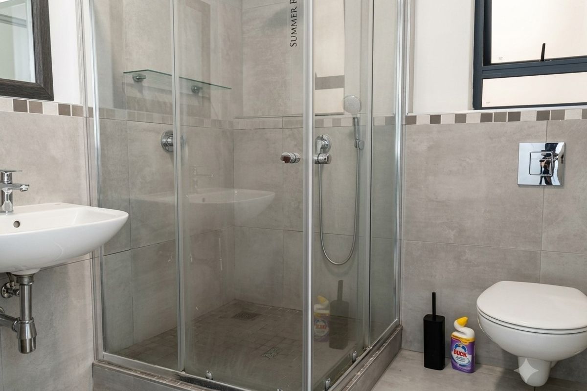 Modern bathroom featuring a glass shower enclosure, wall-mounted toilet, and pedestal basin with neutral grey tiling throughout.