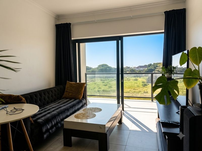 A bright living room featuring large sliding glass doors that open onto a balcony with scenic green views and tiled flooring throughout.