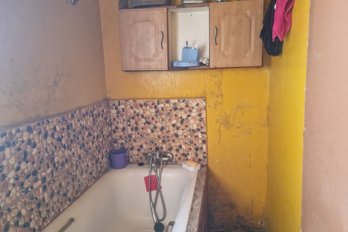 A dated bathroom featuring a bathtub with pebble-style tile splashback and wall-mounted wooden cabinetry, requiring significant cosmetic renovation and repair.