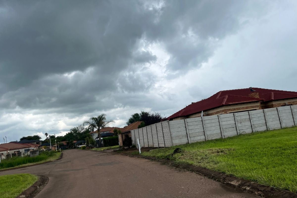 A street view of a residential area featuring paved roads, concrete boundary walls, and surrounding residential properties under a cloudy sky.