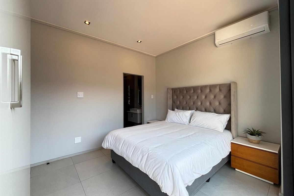 A well-maintained bedroom featuring neutral tiled flooring, recessed ceiling lighting, and a wall-mounted air conditioning unit, with an adjoining en-suite bathroom.