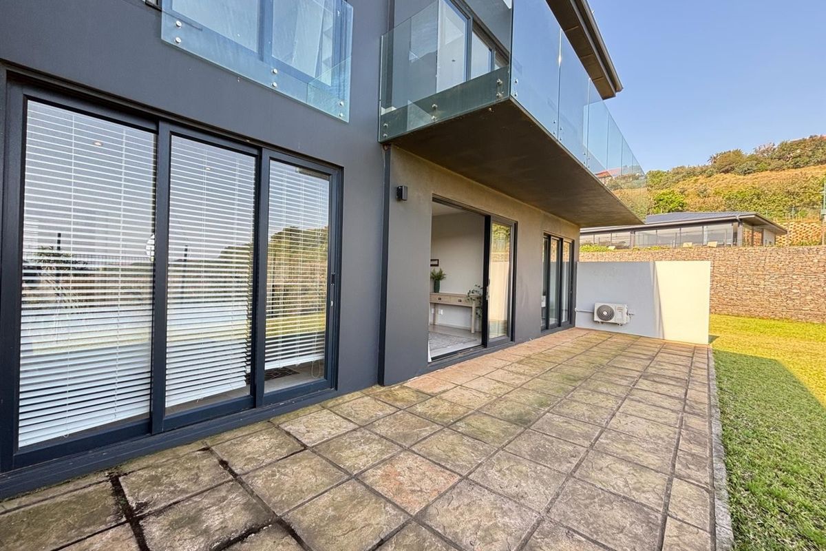 A rear exterior view of the apartment featuring a paved patio area, glass balcony railings, and sliding door access to the interior living space.