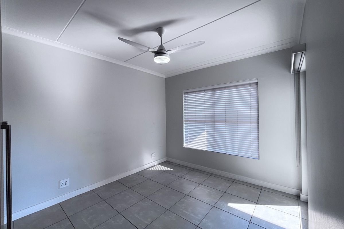 A bright, empty bedroom featuring neutral tiled flooring, a ceiling fan, and venetian blinds, offering a clean and versatile space.