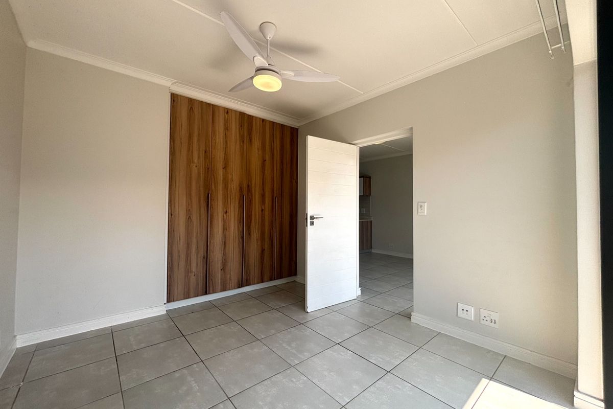 A bright bedroom featuring modern wood-finish built-in cupboards, tiled flooring, and a ceiling fan, with a doorway leading to an adjacent living area.