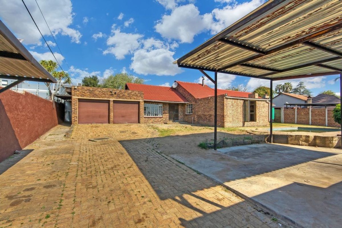 Rear exterior view featuring a double garage, paved driveway, carport structure, and a swimming pool set against a brick house with a tiled roof.