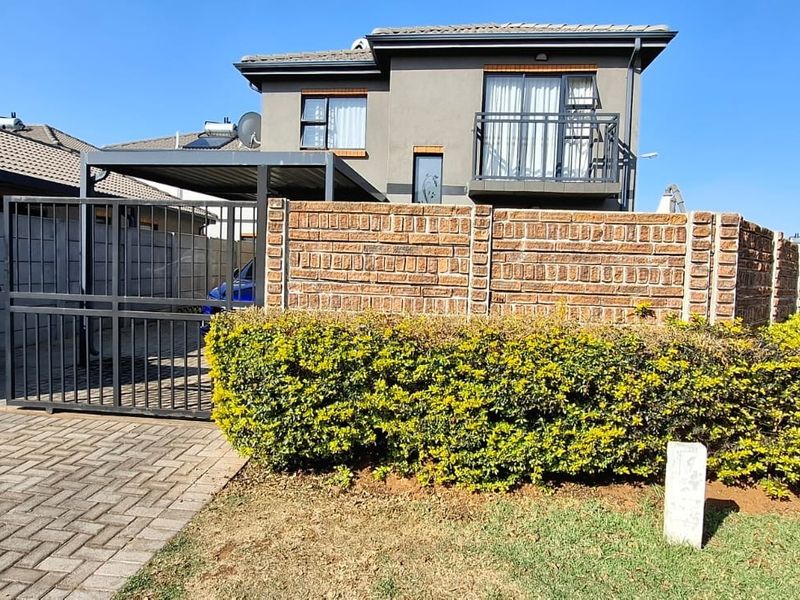 Front view of a modern two-story house featuring a brick boundary wall, metal driveway gate, paved driveway, carport, and an upper-level balcony.