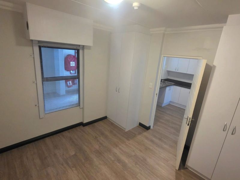 A clean, empty bedroom featuring light-toned laminate flooring, built-in cupboards, and a view into the adjacent kitchen area.