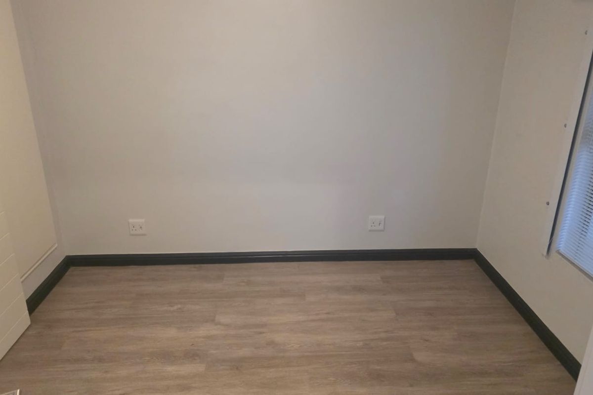 A clean, empty bedroom featuring modern laminate flooring, dark skirting boards, and a window fitted with venetian blinds.