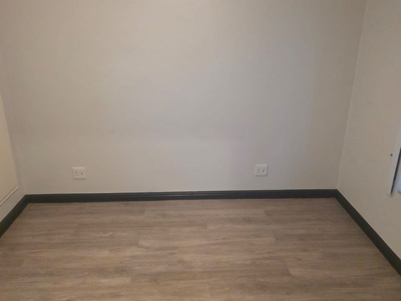 A clean, empty bedroom featuring modern laminate flooring, dark skirting boards, and a window fitted with venetian blinds.