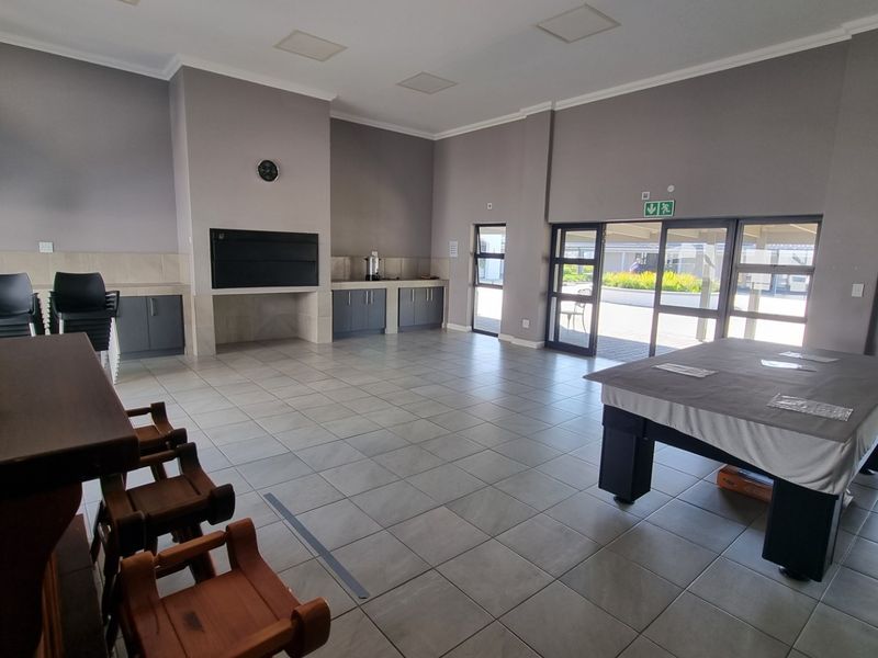 Spacious communal clubhouse area featuring tiled flooring, built-in braai, kitchenette cabinetry, and large glass doors providing access to the outdoor area.