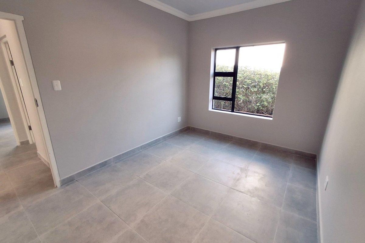 A clean, empty bedroom featuring neutral grey tiled flooring, smooth painted walls, and a modern black-framed window overlooking the garden.