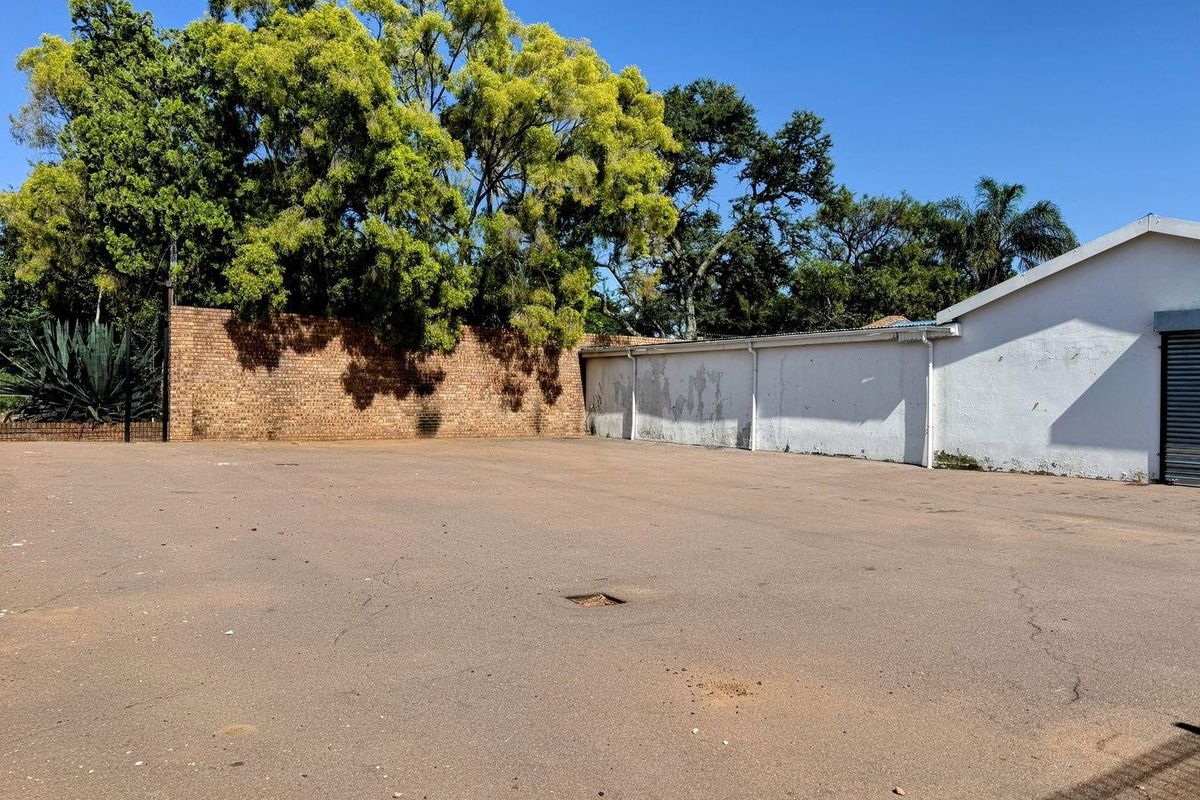 A spacious paved outdoor area featuring a brick boundary wall, mature trees, and a building exterior with a roller shutter door.