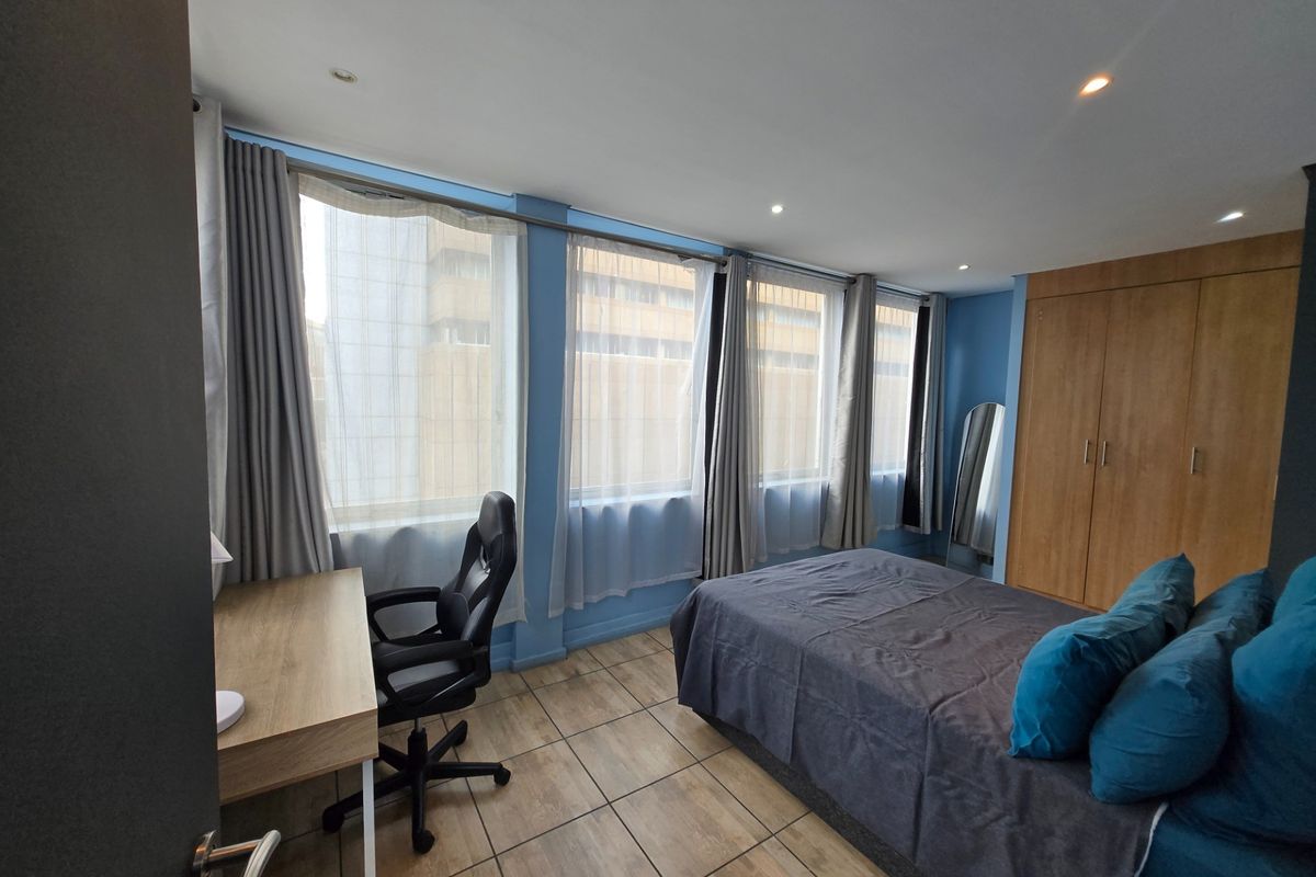 A bedroom featuring built-in wooden wardrobes, tiled flooring, and large windows providing natural light and an urban view.