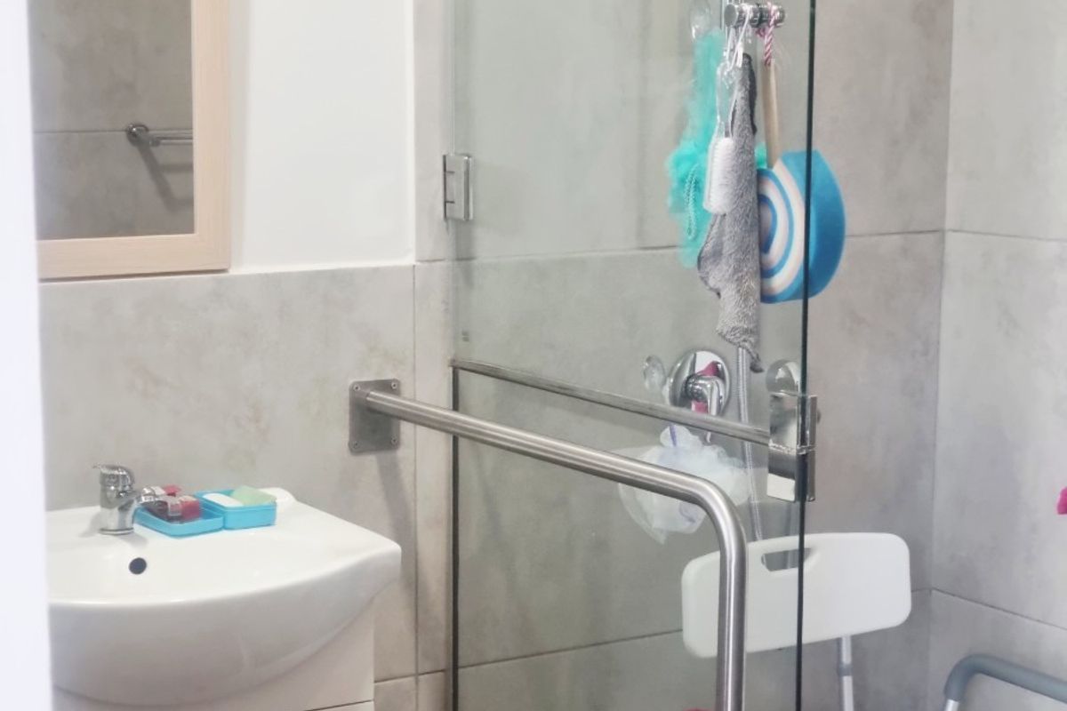 A functional bathroom featuring a wall-mounted vanity, basin, and a glass-enclosed shower with a safety grab rail and tiled finishes.